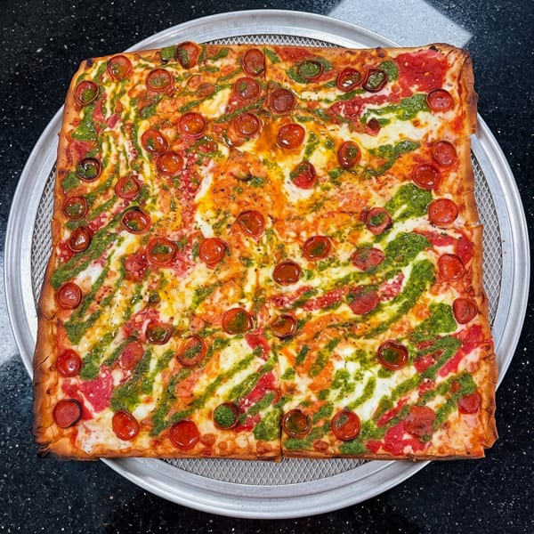 Square pizza with pepperoni, pesto drizzle, and cheese on a round metal pan.