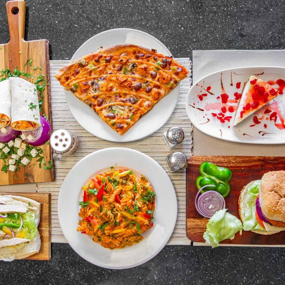 Overhead view of a diverse meal spread including pizza, pasta, wraps, a sandwich, burger, and cheesecake.