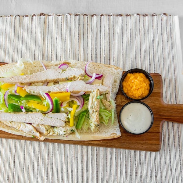 A chicken sub sandwich with lettuce, bell peppers, and red onions, served with two dipping sauces.