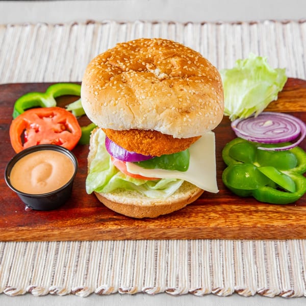 A chicken burger with cheese, lettuce, tomato, onion, and bell pepper on a wooden board.