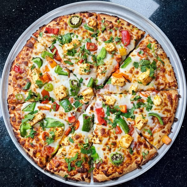 Sliced pizza with melted cheese, red and green peppers, paneer, and fresh cilantro on a metal tray.